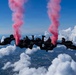 A group of skydivers in black uniforms perform a formation freefall at high altitude, releasing pink/red colored smoke from canisters attached to their gear. The jumpers appear to be members of a military demonstration team, identifiable by 'ARMY' patches visible on their uniforms. They are holding a horizontal linked formation against a backdrop of blue sky, cumulus clouds, and an urban landscape far below.