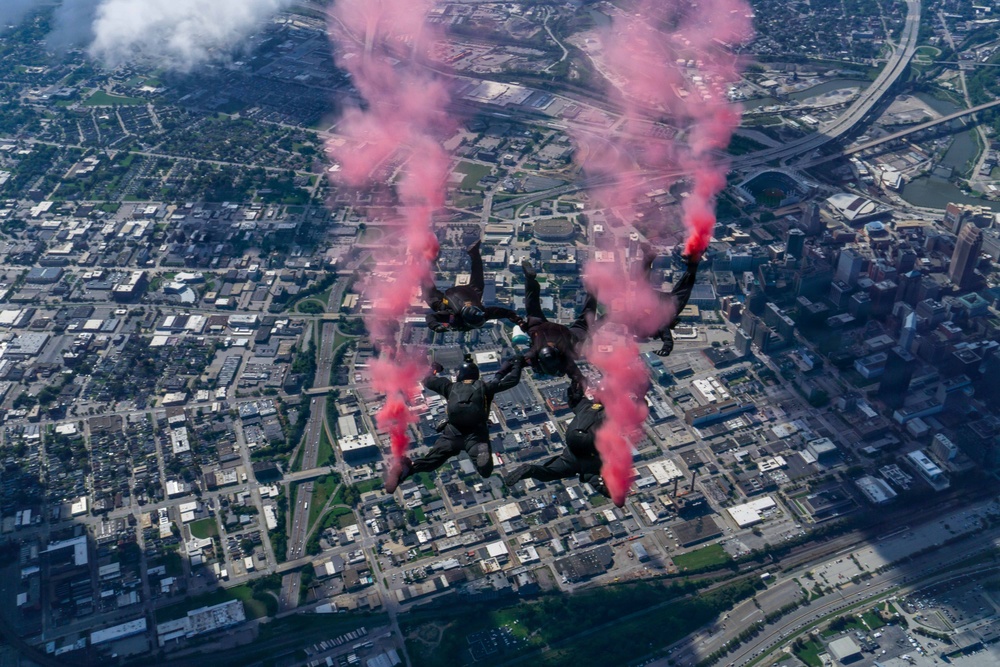 A group of skydivers in black suits are performing a formation freefall over a large urban city, releasing red/pink smoke from handheld or leg-mounted smoke canisters. The aerial perspective reveals a dense metropolitan area with a visible downtown skyline, highway interchanges, and a river or waterway on the right edge. The red smoke trails create dramatic visual streaks against the cityscape below.