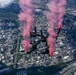 A group of skydivers in black suits are performing a formation freefall over a large urban city, releasing red/pink smoke from handheld or leg-mounted smoke canisters. The aerial perspective reveals a dense metropolitan area with a visible downtown skyline, highway interchanges, and a river or waterway on the right edge. The red smoke trails create dramatic visual streaks against the cityscape below.
