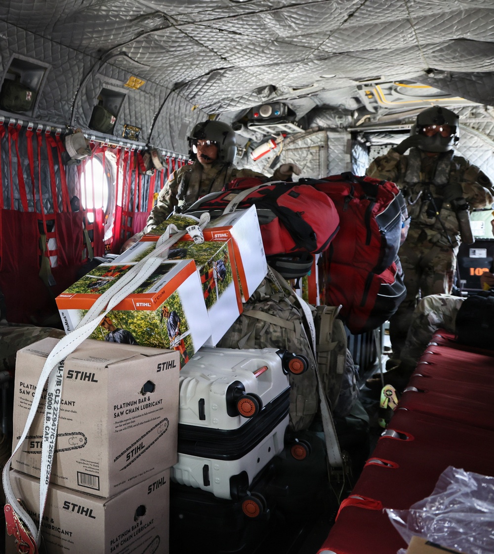 The interior of a military helicopter loaded with cargo including STIHL brand chainsaw equipment and lubricant boxes, a white rolling suitcase, and red duffel bags. Two military personnel wearing flight helmets and camouflage uniforms are visible managing the cargo. This appears to be a disaster relief or humanitarian aid mission given the combination of civilian supplies and military transport.