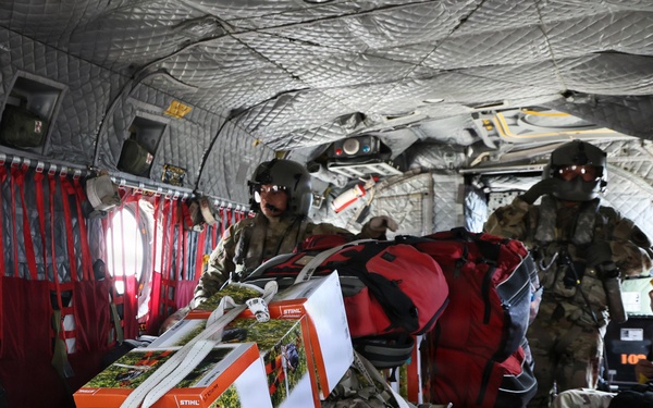 The interior of a military helicopter loaded with cargo including STIHL brand chainsaw equipment and lubricant boxes, a white rolling suitcase, and red duffel bags. Two military personnel wearing flight helmets and camouflage uniforms are visible managing the cargo. This appears to be a disaster relief or humanitarian aid mission given the combination of civilian supplies and military transport.