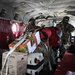 The interior of a military helicopter loaded with cargo including STIHL brand chainsaw equipment and lubricant boxes, a white rolling suitcase, and red duffel bags. Two military personnel wearing flight helmets and camouflage uniforms are visible managing the cargo. This appears to be a disaster relief or humanitarian aid mission given the combination of civilian supplies and military transport.