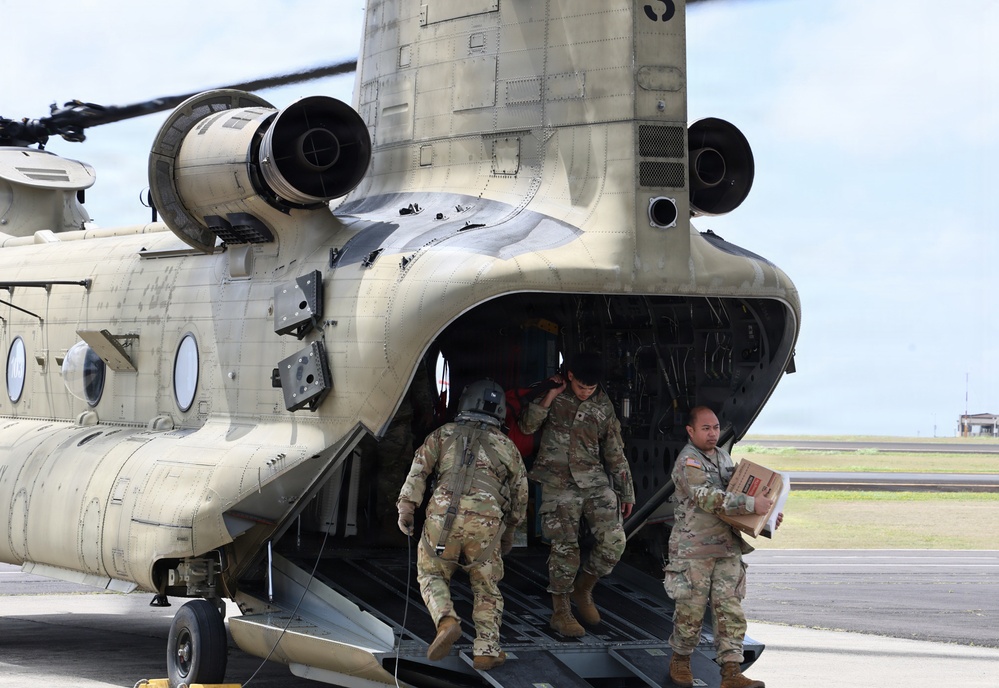 Military personnel are unloading cargo from a CH-47 Chinook helicopter on a tarmac or airfield. At least three soldiers in MultiCam/OCP camouflage uniforms are visible, with one carrying a cardboard box down the rear loading ramp. The helicopter appears to be in a desert tan paint scheme, consistent with U.S. Army aviation assets.