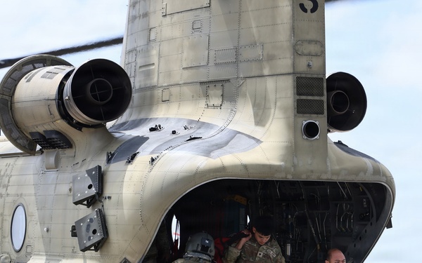 Military personnel are unloading cargo from a CH-47 Chinook helicopter on a tarmac or airfield. At least three soldiers in MultiCam/OCP camouflage uniforms are visible, with one carrying a cardboard box down the rear loading ramp. The helicopter appears to be in a desert tan paint scheme, consistent with U.S. Army aviation assets.