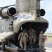 Military personnel are unloading cargo from a CH-47 Chinook helicopter on a tarmac or airfield. At least three soldiers in MultiCam/OCP camouflage uniforms are visible, with one carrying a cardboard box down the rear loading ramp. The helicopter appears to be in a desert tan paint scheme, consistent with U.S. Army aviation assets.