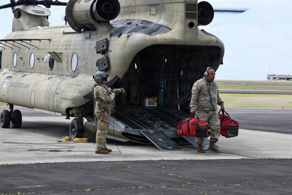 Two U.S. Army soldiers are conducting loading or unloading operations at the rear ramp of a CH-47 Chinook helicopter on a tarmac. One soldier wearing a flight helmet is standing near the ramp while a second soldier carries two red duffel bags away from the aircraft. The helicopter is clearly marked 'United States Army' and appears to be in an operational or training context.