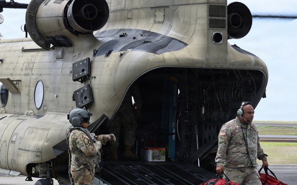 Two U.S. Army soldiers are conducting loading or unloading operations at the rear ramp of a CH-47 Chinook helicopter on a tarmac. One soldier wearing a flight helmet is standing near the ramp while a second soldier carries two red duffel bags away from the aircraft. The helicopter is clearly marked 'United States Army' and appears to be in an operational or training context.