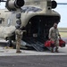 Two U.S. Army soldiers are conducting loading or unloading operations at the rear ramp of a CH-47 Chinook helicopter on a tarmac. One soldier wearing a flight helmet is standing near the ramp while a second soldier carries two red duffel bags away from the aircraft. The helicopter is clearly marked 'United States Army' and appears to be in an operational or training context.
