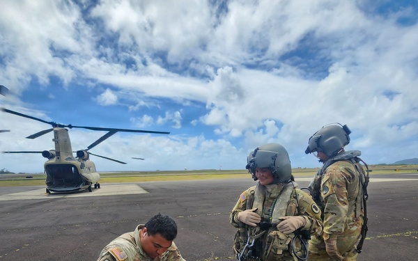 Three U.S. Army soldiers in OCP (Operational Camouflage Pattern) uniforms are on a tarmac or airfield, with a CH-47 Chinook helicopter visible in the background. One soldier is crouching and reviewing documents or paperwork, while two others wearing flight helmets and aviation gear stand nearby. The image is date-stamped September 05, 2026.