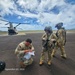 Three U.S. Army soldiers in OCP (Operational Camouflage Pattern) uniforms are on a tarmac or airfield, with a CH-47 Chinook helicopter visible in the background. One soldier is crouching and reviewing documents or paperwork, while two others wearing flight helmets and aviation gear stand nearby. The image is date-stamped September 05, 2026.