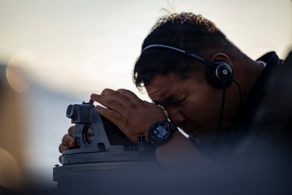 A young man wearing a single-ear headset and a digital watch is looking through a mounted optical sighting instrument, likely a pelorus or bearing circle used for navigation. The image is taken at close range with a shallow depth of field, emphasizing the subject's focused posture. The background suggests an outdoor maritime or elevated environment during low-light conditions such as dawn or dusk.