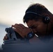 A young man wearing a single-ear headset and a digital watch is looking through a mounted optical sighting instrument, likely a pelorus or bearing circle used for navigation. The image is taken at close range with a shallow depth of field, emphasizing the subject's focused posture. The background suggests an outdoor maritime or elevated environment during low-light conditions such as dawn or dusk.