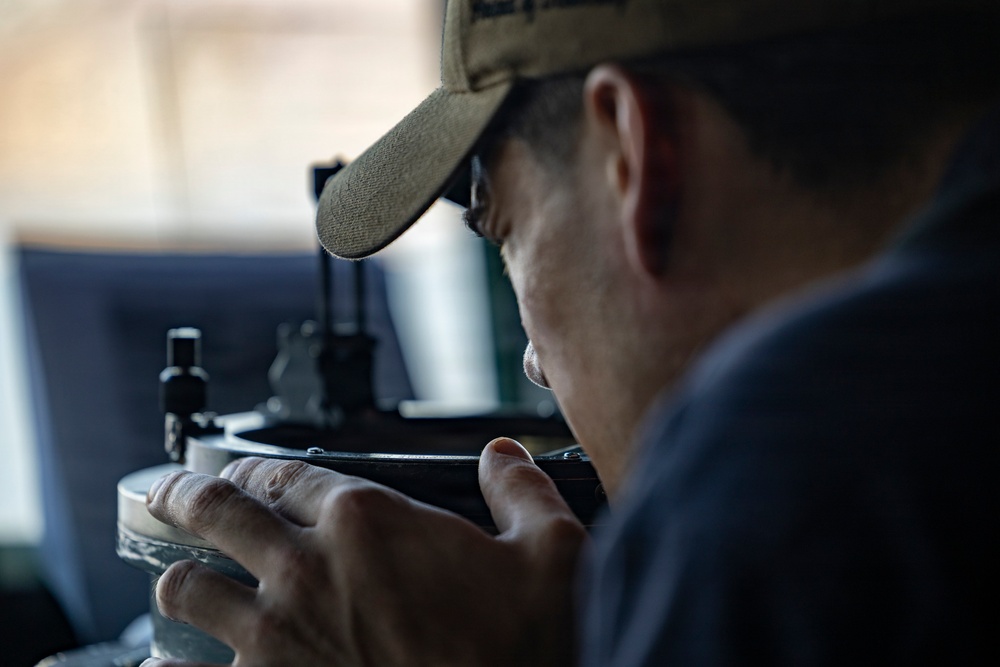 A man wearing a olive/tan baseball cap is closely examining or looking through a precision optical or mechanical instrument. He is holding the device with both hands and appears to be peering through an eyepiece or sighting mechanism. The image is captured in a close-up, shallow depth-of-field style with a blurred background.