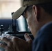 A man wearing a olive/tan baseball cap is closely examining or looking through a precision optical or mechanical instrument. He is holding the device with both hands and appears to be peering through an eyepiece or sighting mechanism. The image is captured in a close-up, shallow depth-of-field style with a blurred background.