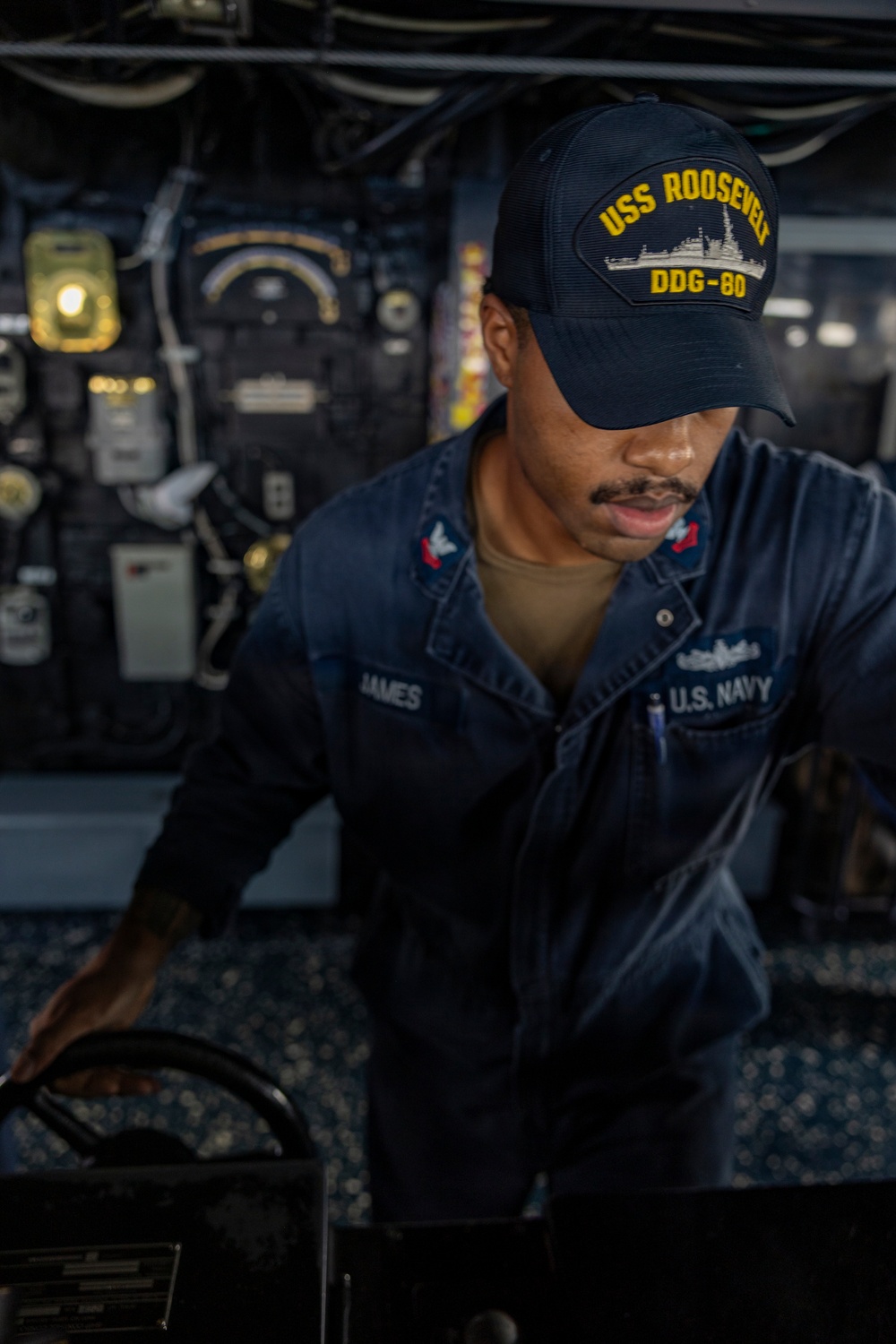A U.S. Navy sailor assigned to USS Roosevelt (DDG-80) is working at a control station aboard the ship. The sailor, identified by name tape as 'JAMES,' is wearing a navy blue coverall uniform and a USS Roosevelt DDG-80 ball cap. The background shows a dimly lit shipboard interior with various gauges, panels, and equipment consistent with a naval vessel's operational spaces.