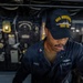 A U.S. Navy sailor assigned to USS Roosevelt (DDG-80) is working at a control station aboard the ship. The sailor, identified by name tape as 'JAMES,' is wearing a navy blue coverall uniform and a USS Roosevelt DDG-80 ball cap. The background shows a dimly lit shipboard interior with various gauges, panels, and equipment consistent with a naval vessel's operational spaces.
