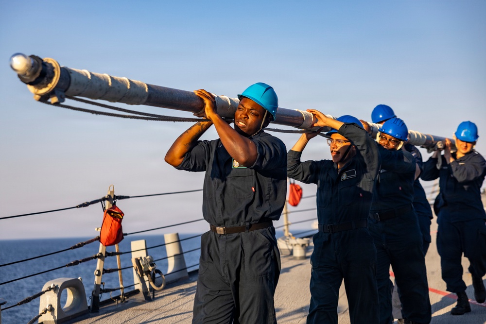 A group of naval personnel in dark blue coveralls and blue hard hats are collectively carrying a large metal pole or spar on the deck of a military vessel at sea. The team appears to be conducting a seamanship or deck evolution exercise, working in coordinated effort. The image is taken during golden hour lighting, suggesting early morning or late afternoon operations.
