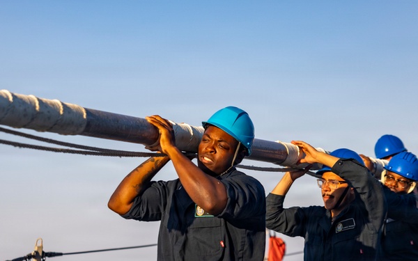 A group of naval personnel in dark blue coveralls and blue hard hats are collectively carrying a large metal pole or spar on the deck of a military vessel at sea. The team appears to be conducting a seamanship or deck evolution exercise, working in coordinated effort. The image is taken during golden hour lighting, suggesting early morning or late afternoon operations.