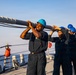 A group of naval personnel in dark blue coveralls and blue hard hats are collectively carrying a large metal pole or spar on the deck of a military vessel at sea. The team appears to be conducting a seamanship or deck evolution exercise, working in coordinated effort. The image is taken during golden hour lighting, suggesting early morning or late afternoon operations.