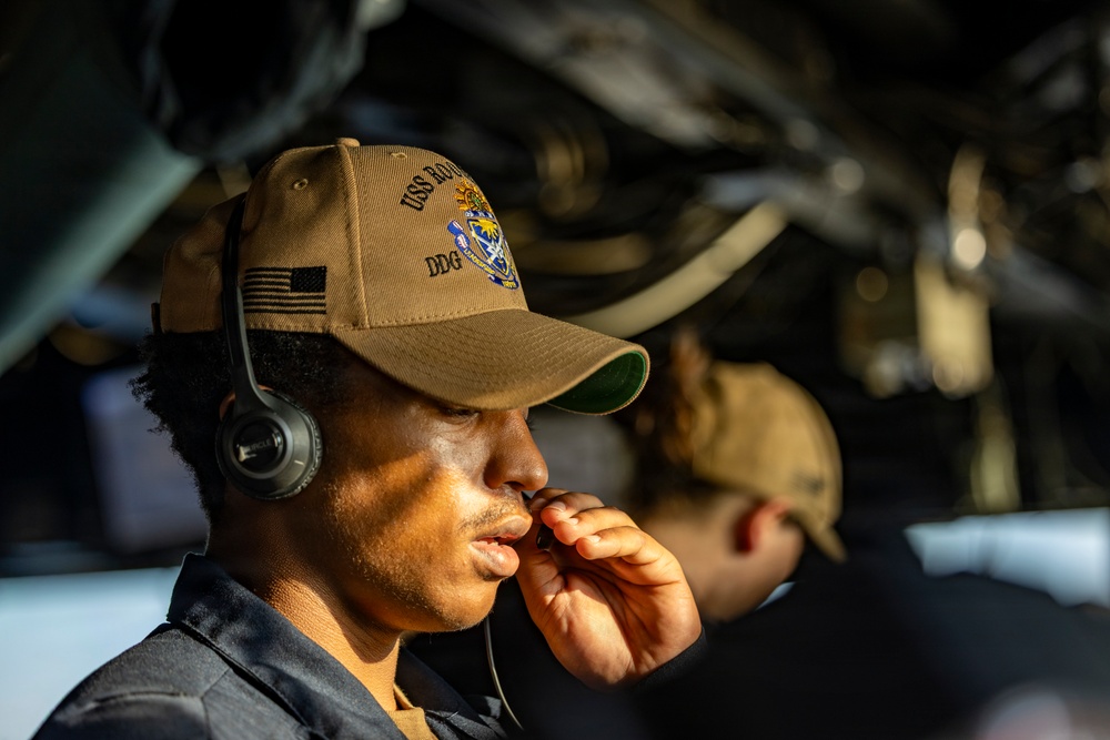 A U.S. Navy sailor wearing a tan ball cap with 'USS' and 'DDG' markings and an American flag patch is shown in a focused, contemplative pose while wearing a communications headset. The sailor appears to be in an operational environment aboard a naval vessel, likely on the bridge or in a combat information center. A second person is visible but out of focus in the background.