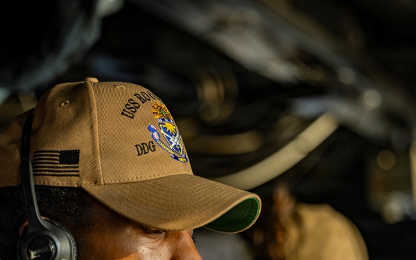 A U.S. Navy sailor wearing a tan ball cap with 'USS' and 'DDG' markings and an American flag patch is shown in a focused, contemplative pose while wearing a communications headset. The sailor appears to be in an operational environment aboard a naval vessel, likely on the bridge or in a combat information center. A second person is visible but out of focus in the background.