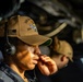 A U.S. Navy sailor wearing a tan ball cap with 'USS' and 'DDG' markings and an American flag patch is shown in a focused, contemplative pose while wearing a communications headset. The sailor appears to be in an operational environment aboard a naval vessel, likely on the bridge or in a combat information center. A second person is visible but out of focus in the background.