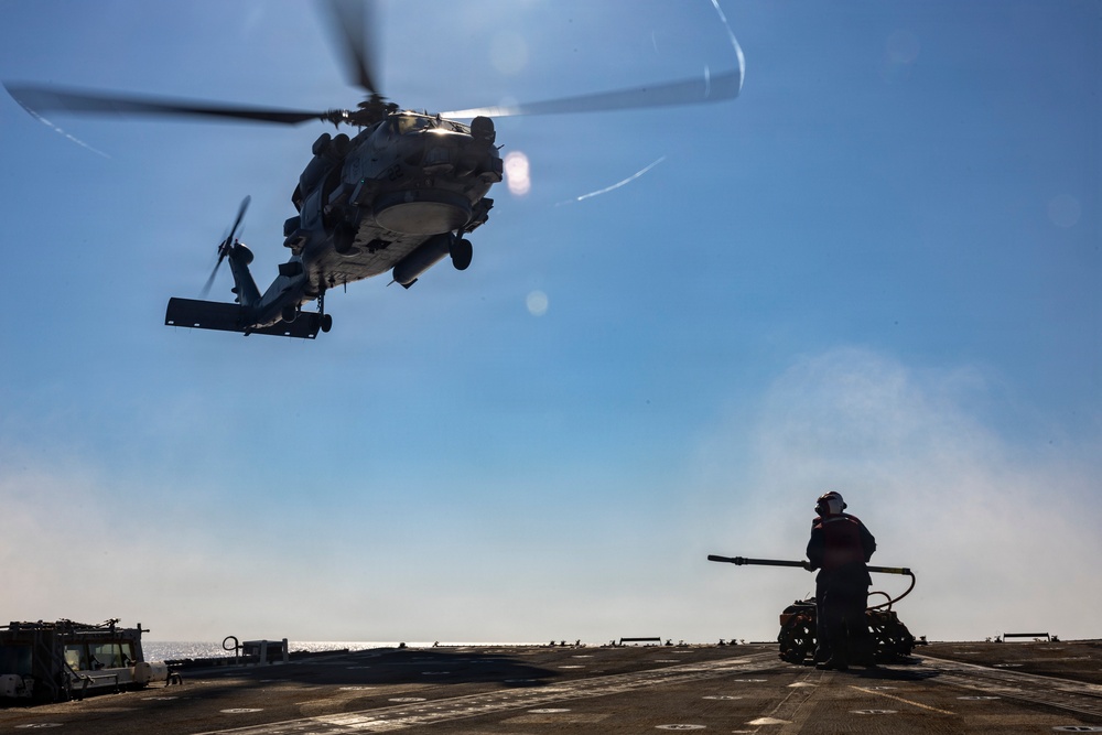 A military helicopter, appearing to be an MH-60 Seahawk variant, hovers low over the flight deck of a naval vessel at sea. A deck crew member in safety gear stands near firefighting or ground support equipment, observing the aircraft. The scene depicts a standard naval flight deck operation under clear, sunny conditions.