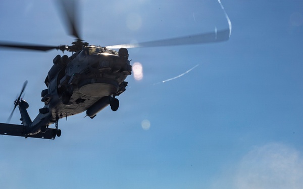 A military helicopter, appearing to be an MH-60 Seahawk variant, hovers low over the flight deck of a naval vessel at sea. A deck crew member in safety gear stands near firefighting or ground support equipment, observing the aircraft. The scene depicts a standard naval flight deck operation under clear, sunny conditions.