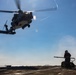 A military helicopter, appearing to be an MH-60 Seahawk variant, hovers low over the flight deck of a naval vessel at sea. A deck crew member in safety gear stands near firefighting or ground support equipment, observing the aircraft. The scene depicts a standard naval flight deck operation under clear, sunny conditions.