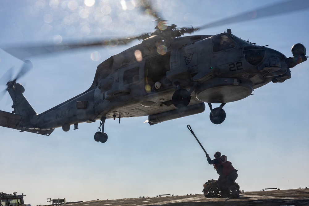 A U.S. Navy helicopter, marked with the number '22' and 'NAVY' on its fuselage, hovers low over a ship's flight deck. A deck crew member is reaching up with a grounding wand toward the aircraft, a standard safety procedure to discharge static electricity before handling. The image is taken from a low angle, emphasizing the scale and proximity of the helicopter to the deck personnel.