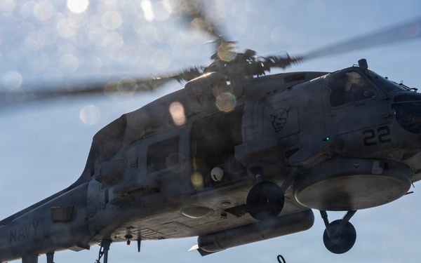 A U.S. Navy helicopter, marked with the number '22' and 'NAVY' on its fuselage, hovers low over a ship's flight deck. A deck crew member is reaching up with a grounding wand toward the aircraft, a standard safety procedure to discharge static electricity before handling. The image is taken from a low angle, emphasizing the scale and proximity of the helicopter to the deck personnel.