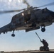 A U.S. Navy helicopter, marked with the number '22' and 'NAVY' on its fuselage, hovers low over a ship's flight deck. A deck crew member is reaching up with a grounding wand toward the aircraft, a standard safety procedure to discharge static electricity before handling. The image is taken from a low angle, emphasizing the scale and proximity of the helicopter to the deck personnel.