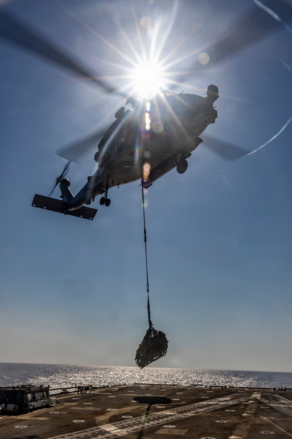 A military helicopter is performing a vertical replenishment (VERTREP) operation over what appears to be the flight deck of a naval vessel at sea. The helicopter is carrying a cargo net loaded with supplies suspended by a sling beneath its fuselage. The image is taken from a low angle looking upward, with the sun creating a dramatic starburst lens flare effect directly behind the aircraft.