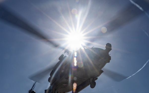 A military helicopter is performing a vertical replenishment (VERTREP) operation over what appears to be the flight deck of a naval vessel at sea. The helicopter is carrying a cargo net loaded with supplies suspended by a sling beneath its fuselage. The image is taken from a low angle looking upward, with the sun creating a dramatic starburst lens flare effect directly behind the aircraft.