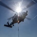 A military helicopter is performing a vertical replenishment (VERTREP) operation over what appears to be the flight deck of a naval vessel at sea. The helicopter is carrying a cargo net loaded with supplies suspended by a sling beneath its fuselage. The image is taken from a low angle looking upward, with the sun creating a dramatic starburst lens flare effect directly behind the aircraft.