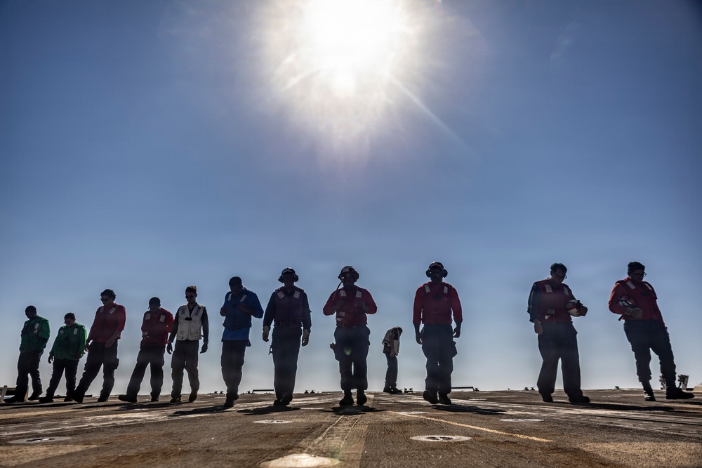 A group of approximately 12 personnel are standing in a line on what appears to be a naval vessel's flight deck, silhouetted against a bright midday sun and clear blue sky. The individuals are wearing various colored vests and safety gear typical of aircraft carrier flight deck crew, suggesting a FOD (Foreign Object Debris) walkdown or inspection procedure. The dramatic backlighting creates strong silhouettes while still revealing color differentiation in their uniforms.
