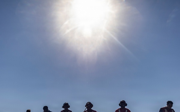 A group of approximately 12 personnel are standing in a line on what appears to be a naval vessel's flight deck, silhouetted against a bright midday sun and clear blue sky. The individuals are wearing various colored vests and safety gear typical of aircraft carrier flight deck crew, suggesting a FOD (Foreign Object Debris) walkdown or inspection procedure. The dramatic backlighting creates strong silhouettes while still revealing color differentiation in their uniforms.
