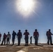 A group of approximately 12 personnel are standing in a line on what appears to be a naval vessel's flight deck, silhouetted against a bright midday sun and clear blue sky. The individuals are wearing various colored vests and safety gear typical of aircraft carrier flight deck crew, suggesting a FOD (Foreign Object Debris) walkdown or inspection procedure. The dramatic backlighting creates strong silhouettes while still revealing color differentiation in their uniforms.