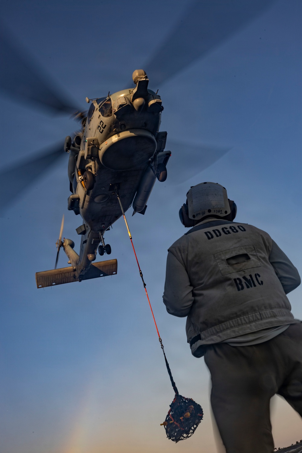A U.S. Navy helicopter, numbered '22', hovers low over a ship's deck during a vertical replenishment (VERTREP) operation. A sailor wearing a vest marked 'DDG80 BMC' guides a cargo net suspended by a red and blue line from the helicopter. The image is taken from a low angle at dusk, creating dramatic lighting conditions.