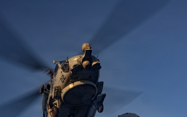 A U.S. Navy helicopter, numbered '22', hovers low over a ship's deck during a vertical replenishment (VERTREP) operation. A sailor wearing a vest marked 'DDG80 BMC' guides a cargo net suspended by a red and blue line from the helicopter. The image is taken from a low angle at dusk, creating dramatic lighting conditions.