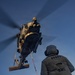A U.S. Navy helicopter, numbered '22', hovers low over a ship's deck during a vertical replenishment (VERTREP) operation. A sailor wearing a vest marked 'DDG80 BMC' guides a cargo net suspended by a red and blue line from the helicopter. The image is taken from a low angle at dusk, creating dramatic lighting conditions.