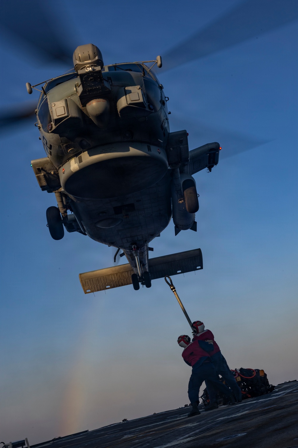 A military helicopter, consistent with an MH-60R Seahawk, hovers low over a naval vessel's flight deck during twilight or early morning hours. A deck crew member wearing a red jersey and cranial helmet is handling a probe or pole connected to the helicopter's underside, likely performing a helicopter in-flight refueling (HIFR) or deck landing assist operation. The dramatic low-angle perspective emphasizes the scale and proximity of the hovering aircraft above the crew member.