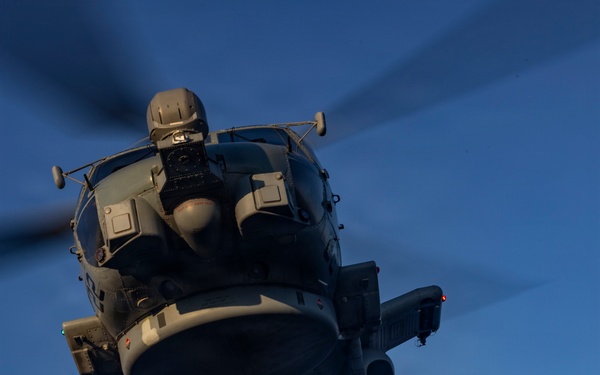 A military helicopter, consistent with an MH-60R Seahawk, hovers low over a naval vessel's flight deck during twilight or early morning hours. A deck crew member wearing a red jersey and cranial helmet is handling a probe or pole connected to the helicopter's underside, likely performing a helicopter in-flight refueling (HIFR) or deck landing assist operation. The dramatic low-angle perspective emphasizes the scale and proximity of the hovering aircraft above the crew member.