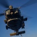A military helicopter, consistent with an MH-60R Seahawk, hovers low over a naval vessel's flight deck during twilight or early morning hours. A deck crew member wearing a red jersey and cranial helmet is handling a probe or pole connected to the helicopter's underside, likely performing a helicopter in-flight refueling (HIFR) or deck landing assist operation. The dramatic low-angle perspective emphasizes the scale and proximity of the hovering aircraft above the crew member.