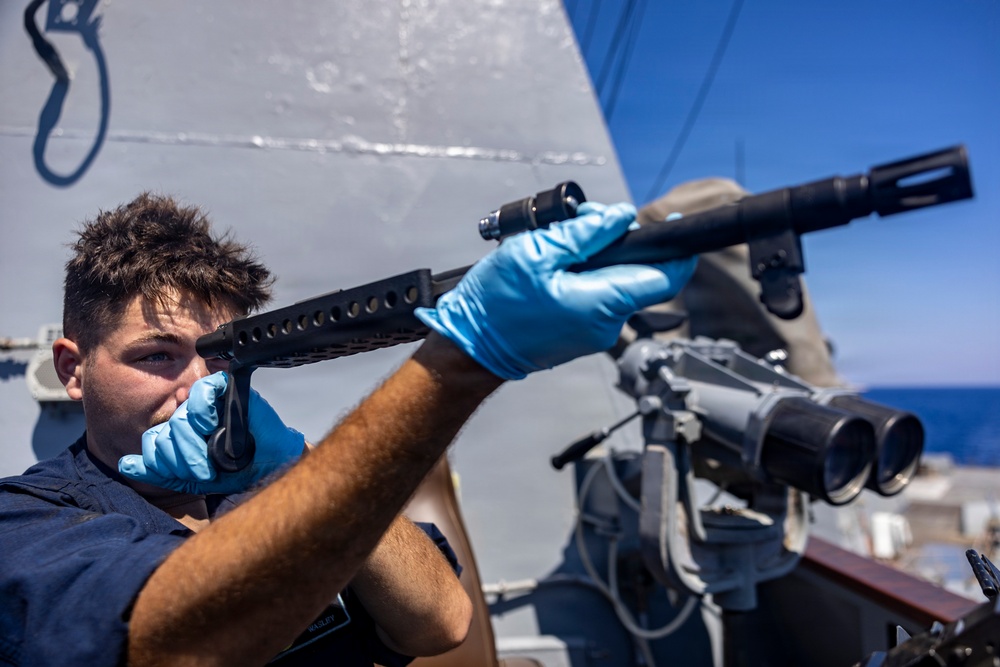 A naval service member wearing blue nitrile gloves inspects or cleans a shotgun barrel on the deck of a military vessel. The sailor is looking through the barrel to check its condition, a standard maintenance procedure. Large binocular optics and ship superstructure are visible in the background.