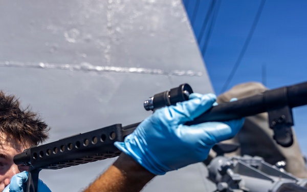 A naval service member wearing blue nitrile gloves inspects or cleans a shotgun barrel on the deck of a military vessel. The sailor is looking through the barrel to check its condition, a standard maintenance procedure. Large binocular optics and ship superstructure are visible in the background.