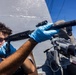 A naval service member wearing blue nitrile gloves inspects or cleans a shotgun barrel on the deck of a military vessel. The sailor is looking through the barrel to check its condition, a standard maintenance procedure. Large binocular optics and ship superstructure are visible in the background.