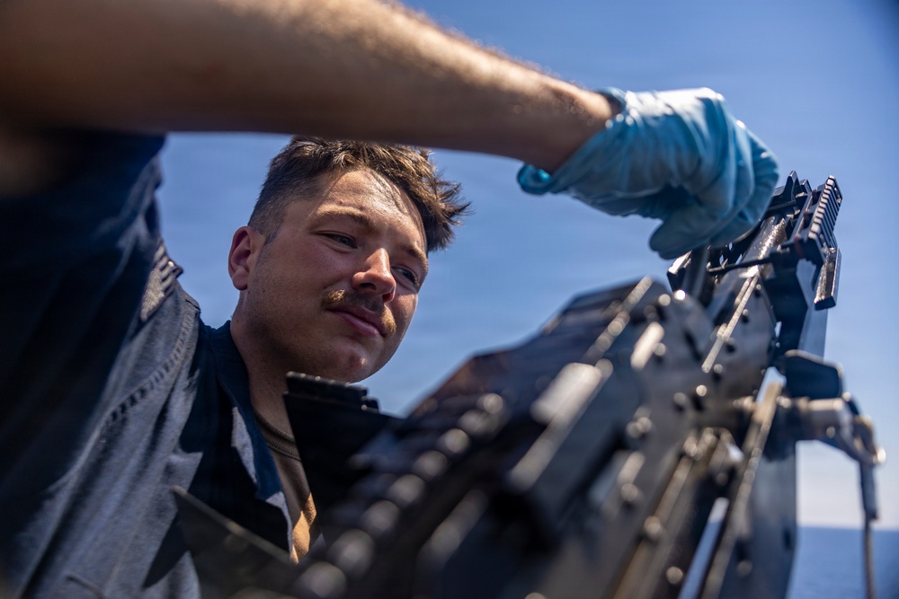 A young male service member is closely inspecting or maintaining a belt-fed machine gun, likely aboard a naval vessel given the open sky and maritime context. A second person wearing a light blue protective glove is assisting from above. The image is taken from a low angle, emphasizing the weapon system and the focused expression of the primary subject.