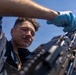 A young male service member is closely inspecting or maintaining a belt-fed machine gun, likely aboard a naval vessel given the open sky and maritime context. A second person wearing a light blue protective glove is assisting from above. The image is taken from a low angle, emphasizing the weapon system and the focused expression of the primary subject.