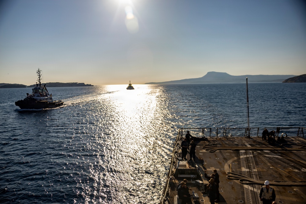 A large naval or military vessel is navigating coastal waters, viewed from its deck, with a tugboat escorting it on the port side. The scene is backlit by a low sun creating a bright glare path across the water, with mountainous coastline visible in the background. Several crew members are visible on the deck, suggesting a port departure or arrival operation.